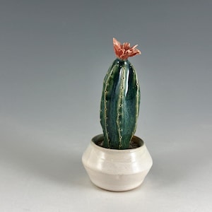 May include: A ceramic cactus with a green and blue glaze and a pink flower on top, sitting in a white ceramic pot.