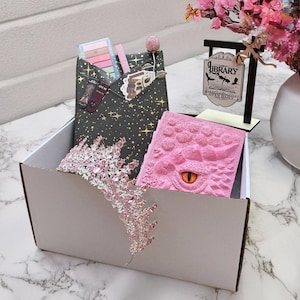 Puede incluir: Una caja de regalo que contiene una tiara rosa, un diario rosa con temática de dragón y un sobre negro con maquillaje y un marcapáginas. También hay un pequeño letrero que dice "Biblioteca" en la caja.