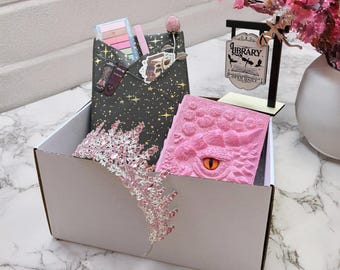 May include: A gift box with a pink tiara, a pink dragon-themed journal, and a black envelope with makeup and a bookmark. A small sign that says "Library" is also in the box.