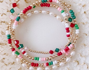 May include: A set of four beaded bracelets in red, green, white, and pink with gold beads and crystal accents. The bracelets are layered on a white surface.