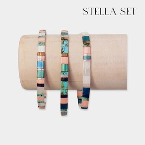 May include: Three colorful beaded bracelets on a wooden display stand. The bracelets are made with turquoise, pink, white, and black beads. The bracelets are labeled "STELLA SET".