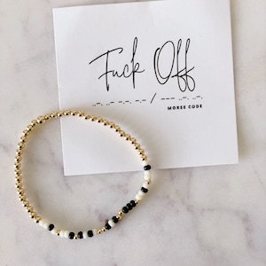 May include: A gold beaded bracelet with black and white beads spelling out "F**k Off" in Morse code. The bracelet is on a white card with the phrase "F**k Off" written in cursive and the Morse code translation below.