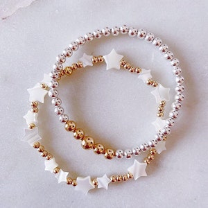 May include: Two bracelets with white star-shaped beads. One bracelet has gold beads and the other has silver beads.