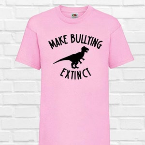 May include: A pale pink t-shirt featuring the words "MAKE BULLYING EXTINCT" in black, with a black dinosaur silhouette in between. The shirt has short sleeves and is made of a soft fabric.