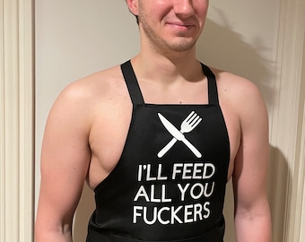 May include: Black apron with white text that reads "I'LL FEED ALL YOU FUCKERS". The apron has a crossed knife and fork graphic. The apron is worn by a person.