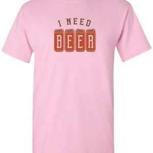 May include: A light pink t-shirt with a crew neck. The shirt features the text "I NEED BEER" printed in brown. The word "BEER" is displayed in a block letter style, with each letter inside a brown beer can.