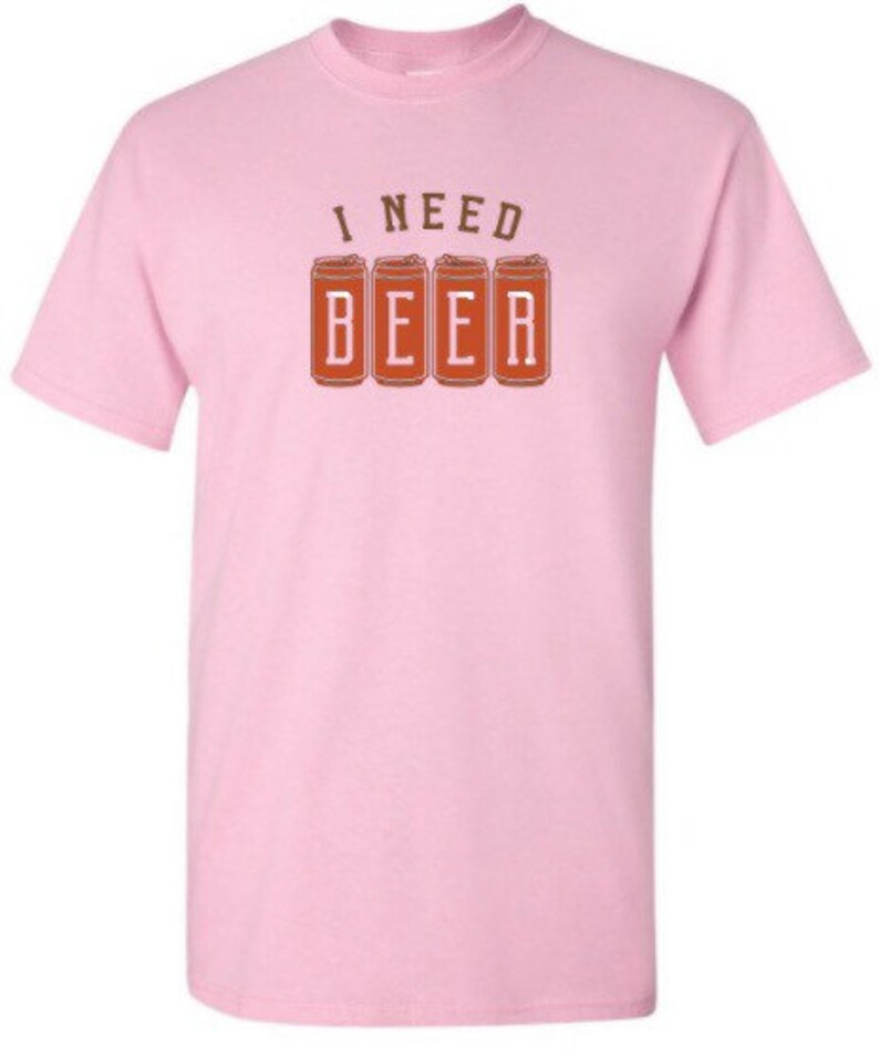 May include: A light pink t-shirt with a crew neck. The shirt features the text "I NEED BEER" printed in brown. The word "BEER" is displayed in a block letter style, with each letter inside a brown beer can.