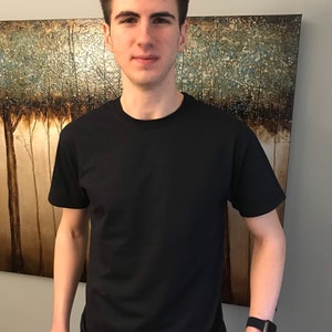 May include: A black crew neck t-shirt. The short-sleeved shirt is made of a soft material. The person is wearing a black smartwatch. The background features a painting with a tree theme.