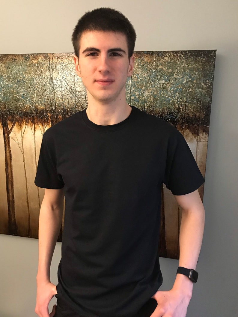 May include: A black crew neck t-shirt. The short-sleeved shirt is made of a soft material. The person is wearing a black smartwatch. The background features a painting with a tree theme.