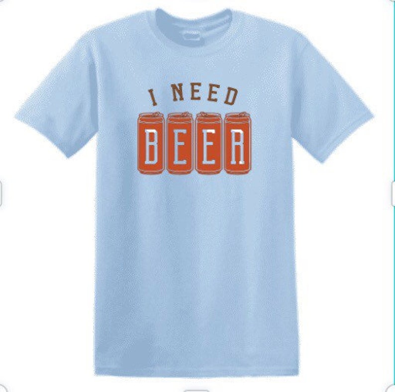 May include: Light blue t-shirt with the phrase "I NEED BEER" printed on the front. The text "I NEED" is in a brown font, and the word "BEER" is displayed in a block letter style with four beer can graphics.
