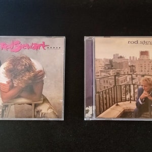 Puede incluir: Dos estuches de discos compactos con álbumes de Rod Stewart. El primer estuche presenta una foto de Rod Stewart con la cabeza en las manos. El título del álbum es "Out of Order". El segundo estuche presenta una foto de Rod Stewart sentado en una azotea con una vista de la ciudad al fondo. El título del álbum es "When We Were The New Boys".
