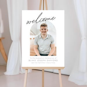 Puede incluir: Un cartel de bienvenida blanco con un arco negro y el texto "welcome" en letra cursiva. Una foto de un joven con una camisa gris claro está en el arco. El texto debajo de la foto dice "To the Graduation Celebration of BLAKE JOSEPH DAFFORD Lake View High School Class of 2023".
