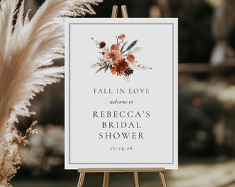 Può includere: Un cartello su un cavalletto dorato con la scritta "FALL IN LOVE welcome to REBECCA'S BRIDAL SHOWER 10.24.26". Il cartello ha una composizione floreale in alto con fiori rossi, arancioni e bianchi. Lo sfondo è sfocato.