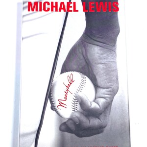May include: A book cover titled Moneyball: The Art of Winning an Unfair Game by Michael Lewis. The cover features a black and white photograph of a hand holding a baseball, with the author's name and the book title printed in bold red text.