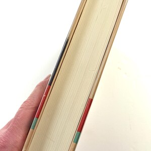 Puede incluir: Un libro de tapa dura con lomo y p&aacute;ginas beige. El lomo tiene detalles en rojo, negro y verde. Las p&aacute;ginas del libro son de color beige claro. El libro est&aacute; sostenido en la mano de una persona.