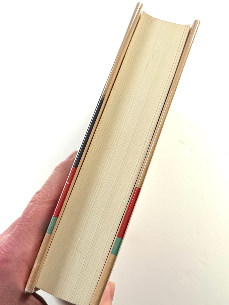 Puede incluir: Un libro de tapa dura con lomo y p&aacute;ginas beige. El lomo tiene detalles en rojo, negro y verde. Las p&aacute;ginas del libro son de color beige claro. El libro est&aacute; sostenido en la mano de una persona.