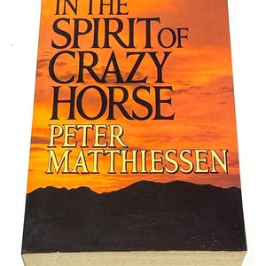 May include: A hardcover book titled "IN THE SPIRIT OF CRAZY HORSE" by Peter Matthiessen. The cover features a sunset-colored sky over a dark mountain range. The title and author's name are in bold, black and gold lettering.
