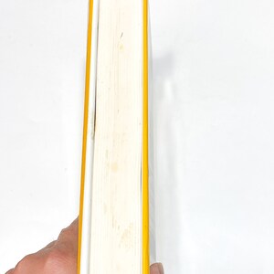 May include: A white book with a yellow spine and blue edges. The book is held in a hand.