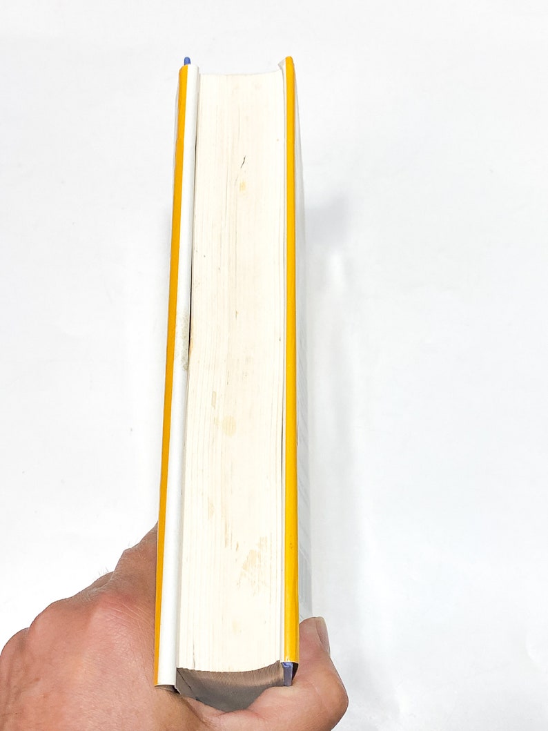 May include: A white book with a yellow spine and blue edges. The book is held in a hand.