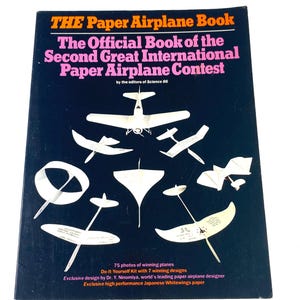 May include: A black book titled The Paper Airplane Book: The Official Book of the Second Great International Paper Airplane Contest by the editors of Science 86. The cover features white illustrations of various paper airplane designs.
