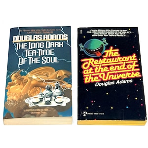 May include: Two paperback books by Douglas Adams are displayed. The book on the left is titled "The Long Dark Tea-Time of the Soul" with a blue cover and gold accents. The book on the right is titled "The Restaurant at the End of the Universe" with a black cover and colorful text.