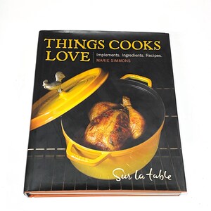 May include: A cookbook titled "Things Cooks Love" by Marie Simmons. The cover features a yellow dutch oven with a rooster finial on the lid. A roasted chicken is inside the pot. The text "Sur la Table" is printed on the bottom of the cover.