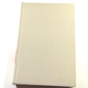 Puede incluir: Un libro de tapa dura con una cubierta de lino beige. El libro est&aacute; cerrado y ligeramente inclinado, revelando un vistazo del lomo con detalles en rojo y azul. La cubierta tiene una textura sutil.