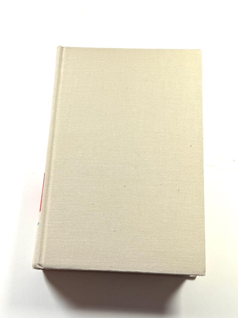 Puede incluir: Un libro de tapa dura con una cubierta de lino beige. El libro est&aacute; cerrado y ligeramente inclinado, revelando un vistazo del lomo con detalles en rojo y azul. La cubierta tiene una textura sutil.