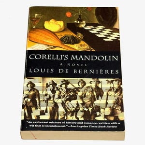 May include: A hardcover book titled "Corelli's Mandolin" by Louis de Bernières. The cover features a still life with a mandolin, a glass of wine, and a checkerboard. The book has a black spine with white text and a quote from the Los Angeles Times.