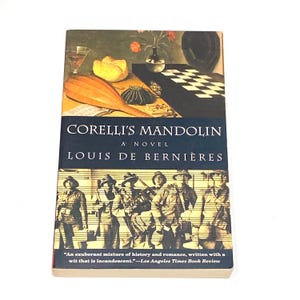 May include: A hardcover book titled "Corelli's Mandolin" by Louis de Bernières. The cover features a painting with a mandolin, flowers, and a chessboard. The bottom of the cover has a black and white image of soldiers. The book has a quote from the Los Angeles Times Book Review.