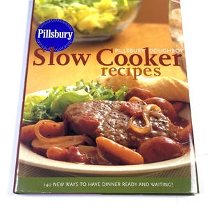 May include: A Pillsbury Slow Cooker recipes cookbook with a blue logo, featuring a photograph of a cooked meat dish with carrots and onions on a plate, and the text 140 NEW WAYS TO HAVE DINNER READY AND WAITING! printed on a green bottom border.