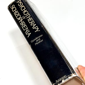 A person is holding a black book titled "PSYCHOTHERAPY OF SCHIZOPHERIA" by Guldenson and Mosher.