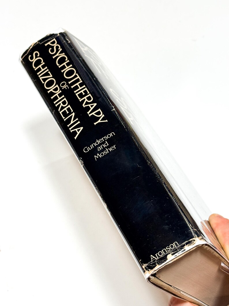 A person is holding a black book titled "PSYCHOTHERAPY OF SCHIZOPHERIA" by Guldenson and Mosher.