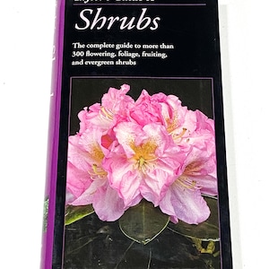 May include: A hardcover book titled "Taylor's Guide to Shrubs." The cover features a vibrant image of pink flowers with yellow centers against a dark background. The book's spine is purple.