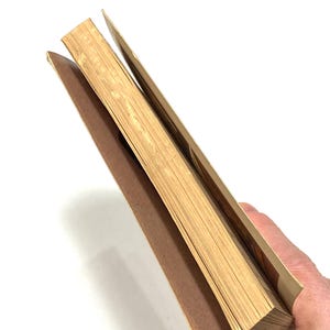 A hand is holding a stack of brown paper or cardboard, with the pages of the book visible.