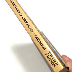 A person's hand holding a cookbook titled "Third Edition" by Ghirardelli, which appears to be a chocolate recipe book.