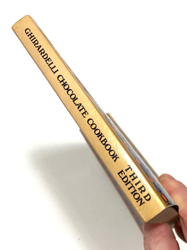 A person's hand holding a cookbook titled "Third Edition" by Ghirardelli, which appears to be a chocolate recipe book.