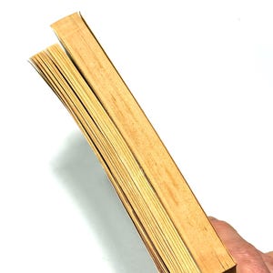 May include: Two vintage books with aged, yellowed pages are held against a white background. The edges of the pages are visible, showing the texture and age of the paper. The covers are a light brown color.