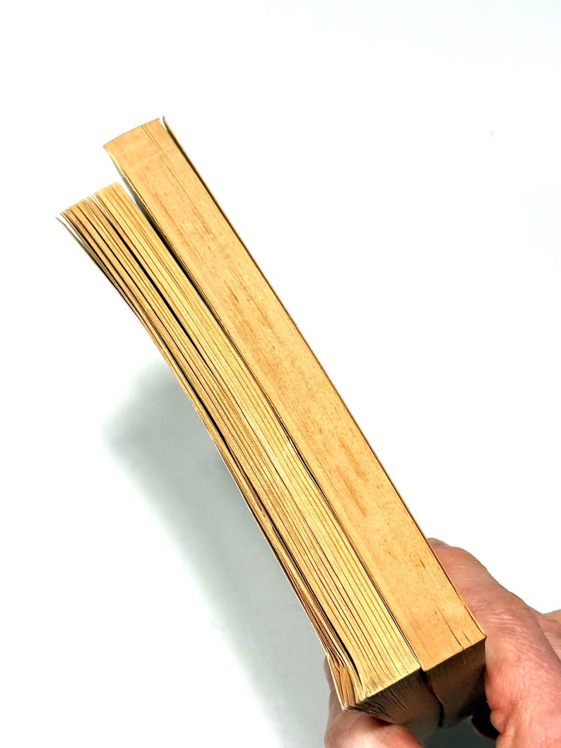 May include: Two vintage books with aged, yellowed pages are held against a white background. The edges of the pages are visible, showing the texture and age of the paper. The covers are a light brown color.