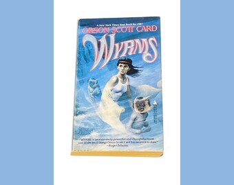 May include: A book cover with a blue and white background. The title "WYRMS" is in red and white lettering. The author's name, "Orson Scott Card", is printed in black lettering. The book cover features a woman with dark hair and a white tank top standing in front of a blue and white background. The woman is holding a white cloth in her hands. There are two small, white, furry creatures in the background.