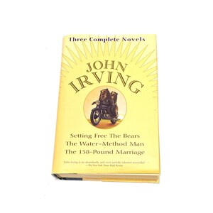 May include: A yellow book cover with a sunburst design. The title is "Three Complete Novels" and the author is "John Irving". The cover features an illustration of two bears riding a motorcycle. The book title is "Setting Free The Bears", "The Water-Method Man", and "The 158-Pound Marriage".