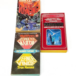 May include: A stack of three science fiction paperback books. The top book is red with gold lettering and the title "TIMESCAPE" by George R.R. Martin and Lisa Tuttle. The middle book is red with gold lettering and the title "Nightflyers" by George R.R. Martin. The bottom book is black with yellow lettering and the title "True Names" by Vernor Vinge.
