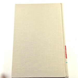 Puede incluir: Un libro de tapa dura con una cubierta de lino beige claro. El lomo tiene marcas rojas y azules. El libro es rectangular y parece cerrado. La cubierta tiene una textura sutil.