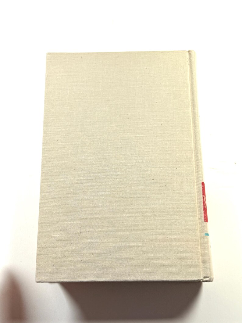 Puede incluir: Un libro de tapa dura con una cubierta de lino beige claro. El lomo tiene marcas rojas y azules. El libro es rectangular y parece cerrado. La cubierta tiene una textura sutil.