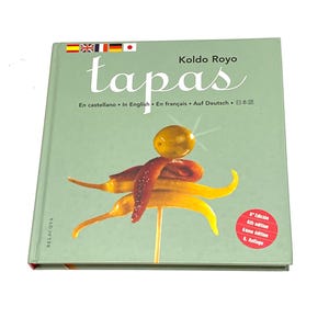 May include: A cookbook titled "tapas" by Koldo Royo, featuring a light green cover with the word "tapas" in large white letters. The cover also displays a skewer of tapas with red, yellow, and brown peppers and an olive.