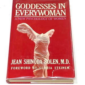 May include: A hardcover book titled "Goddesses in Everywoman" with a red cover. The book features a white statue illustration and text that reads "A New Psychology of Women" and the author's name, Jean Shinoda Bolen, M.D.