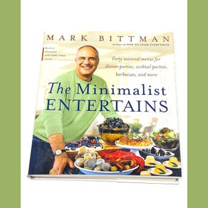 May include: A cookbook titled "The Minimalist Entertains" by Mark Bittman, author of "How to Cook Everything". The cover features a photo of the author standing in front of a table with a variety of food, including seafood, pasta, and desserts.