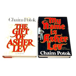 May include: Two hardcover books by Chaim Potok are displayed. The book on the left is titled "The Gift of Asher Lev" with red and gold lettering. The book on the right is titled "My Name is Asher Lev" with red and yellow lettering.