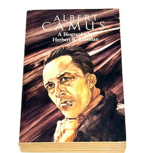 May include: A hardcover biography of Albert Camus by Herbert R. Lottman. The book cover features a portrait of Camus with a cigarette, set against a dark brown and maroon background. The title is in white capital letters.