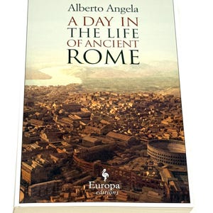 May include: A hardcover book titled "A Day in the Life of Ancient Rome" by Alberto Angela. The cover features an aerial view of ancient Rome with the Europa editions logo. The book has a white spine and pages.